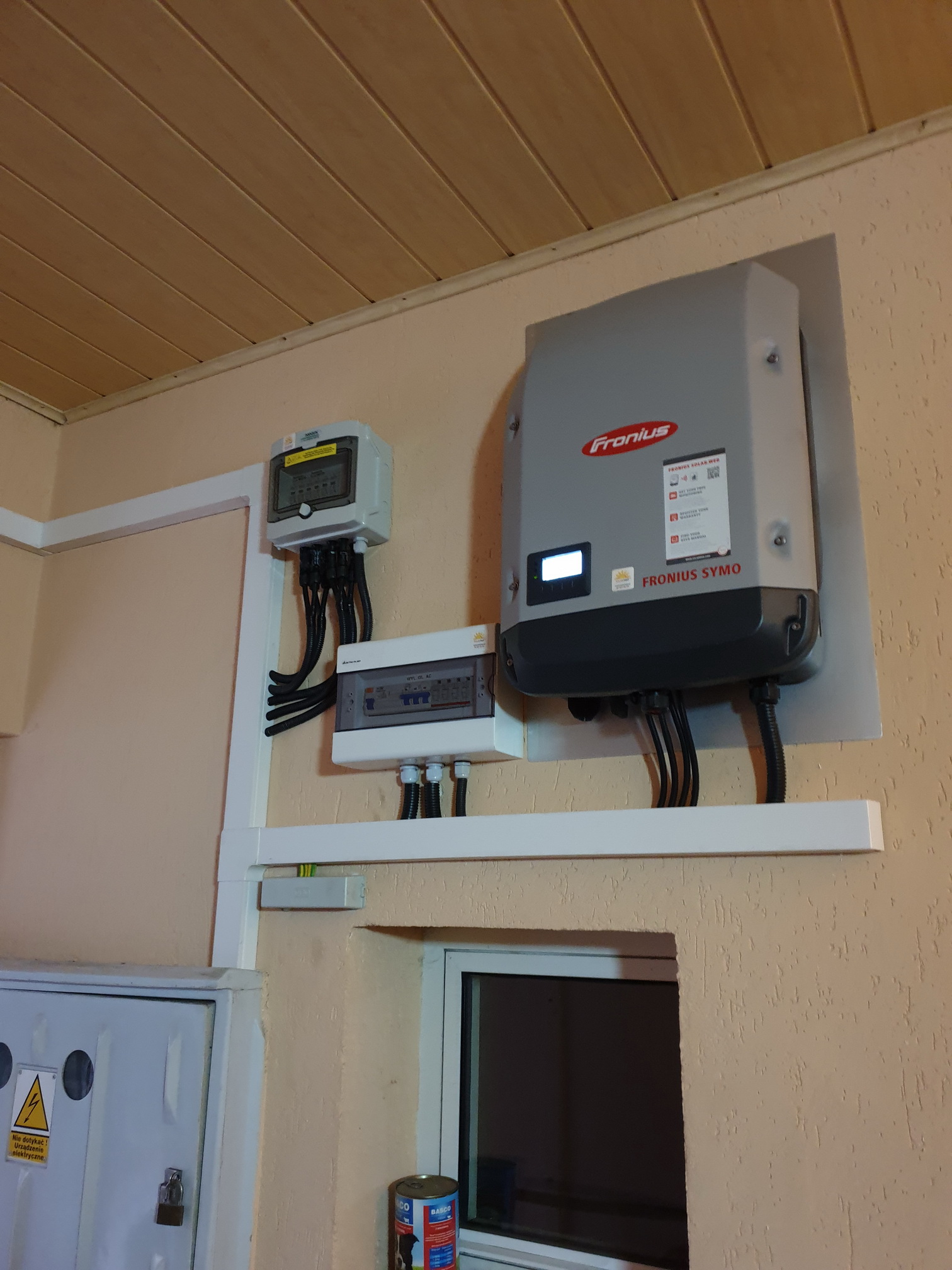 Szary inwerter Fronius SYMO zamontowany na ścianie obok skrzynki z bezpiecznikami i licznika energii elektrycznej, z widocznymi przewodami w peszlach.