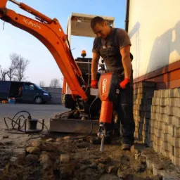 Pracownik używa młota Hilti TE 3000-AVR obok minikoparki Kubota, przygotowując wykop pod chodnik z kostki brukowej. Widoczny kabel zasilający.