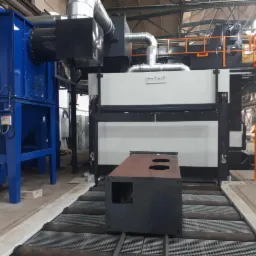 Element stalowy na taśmie transportowej przed automatyczną kabiną do śrutowania, z widocznym niebieskim separatorem pyłu i pomarańczową drabinką serwisową, w hali przemysłowej.