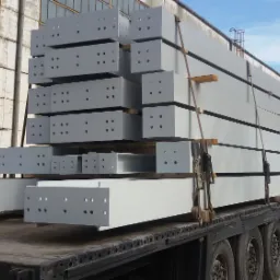 Transport belek stalowych z otworami, zabezpieczonych pasami na naczepie ciężarówki, z widocznym fragmentem starej hali w tle.