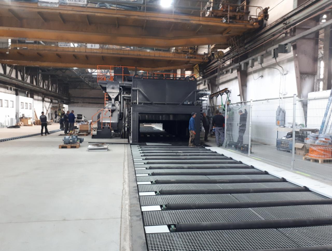 Wnętrze hali przemysłowej z linią transportową prowadzącą do dużej maszyny do obróbki powierzchniowej, prawdopodobnie przed lub po procesie, z grupą osób w tle.