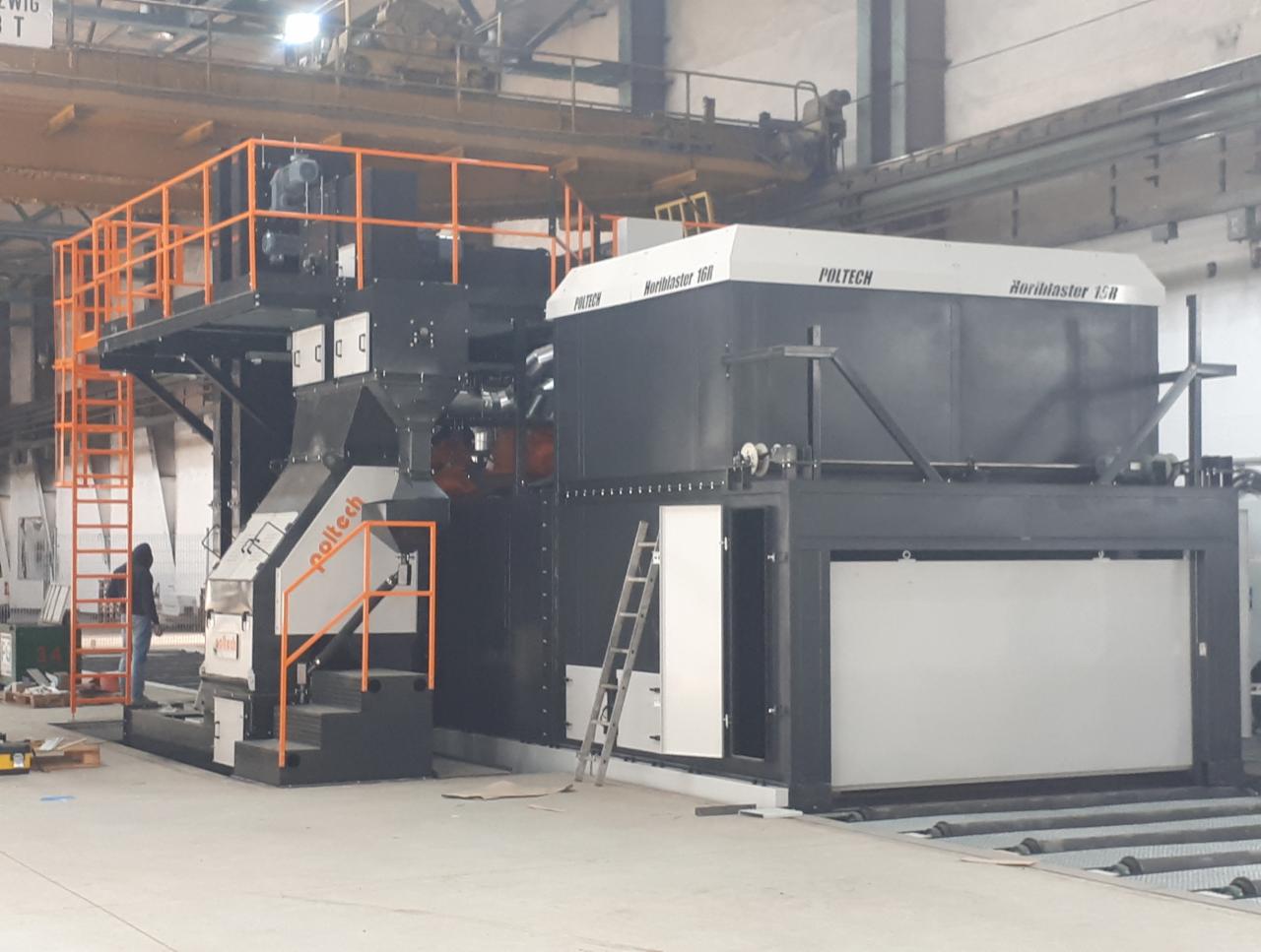 Duża, przemysłowa maszyna do obróbki powierzchni Horiblaster 16R marki Poltech, z pomostem i schodami pomalowanymi na pomarańczowo, ustawiona w hali produkcyjnej, z widoczną osobą w oddali.