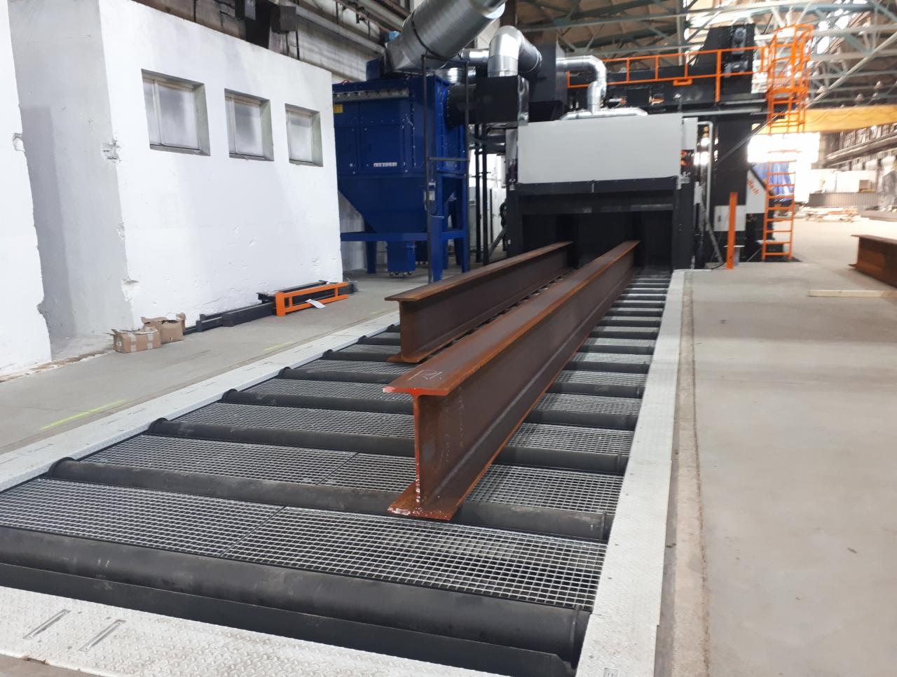 Dwie zardzewiałe stalowe belki H na podajniku rolkowym w hali produkcyjnej, przed automatyczną maszyną do obróbki strumieniowo-ściernej.