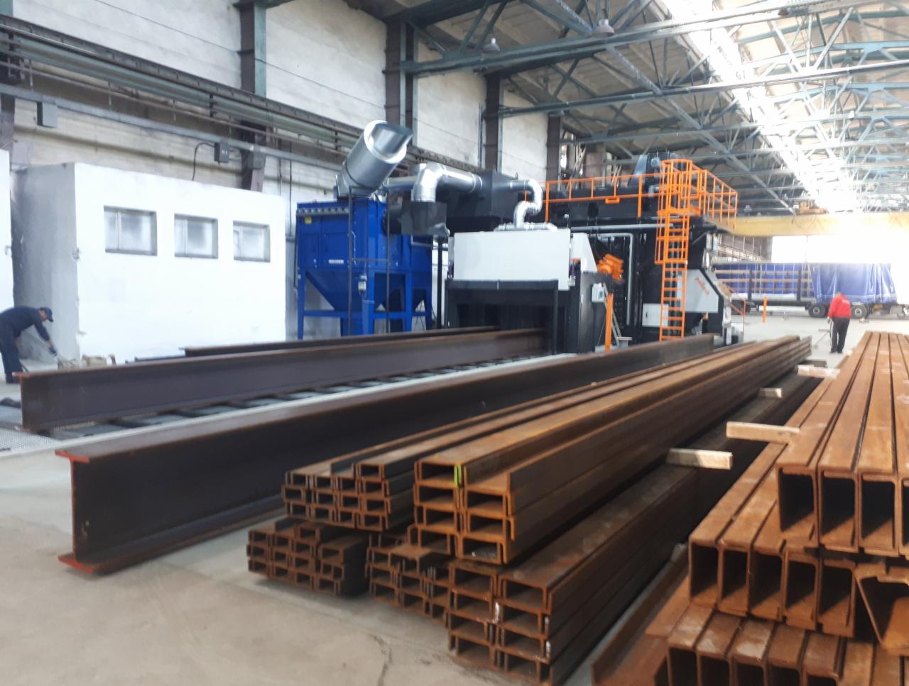 Stalowe profile różnego kształtu i wielkości ułożone na podłodze hali produkcyjnej, w tle maszyna do obróbki strumieniowo-ściernej i pracownik w kombinezonie.