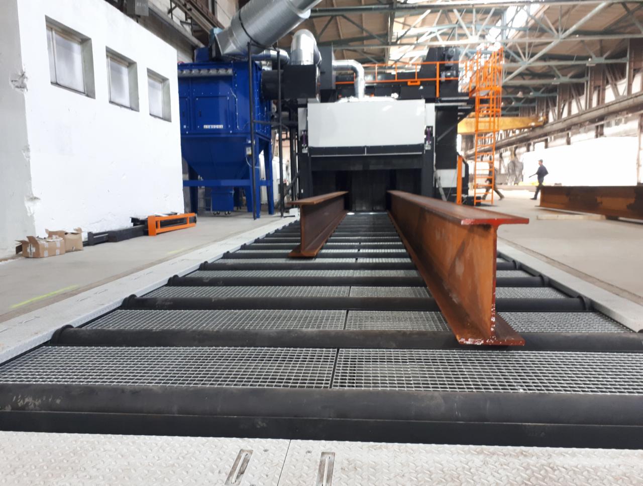 Dwa stalowe profile H na metalowej platformie rolkowej w hali produkcyjnej przed niebieską maszyną do obróbki strumieniowo-ściernej, widoczne ślady korozji na profilach.