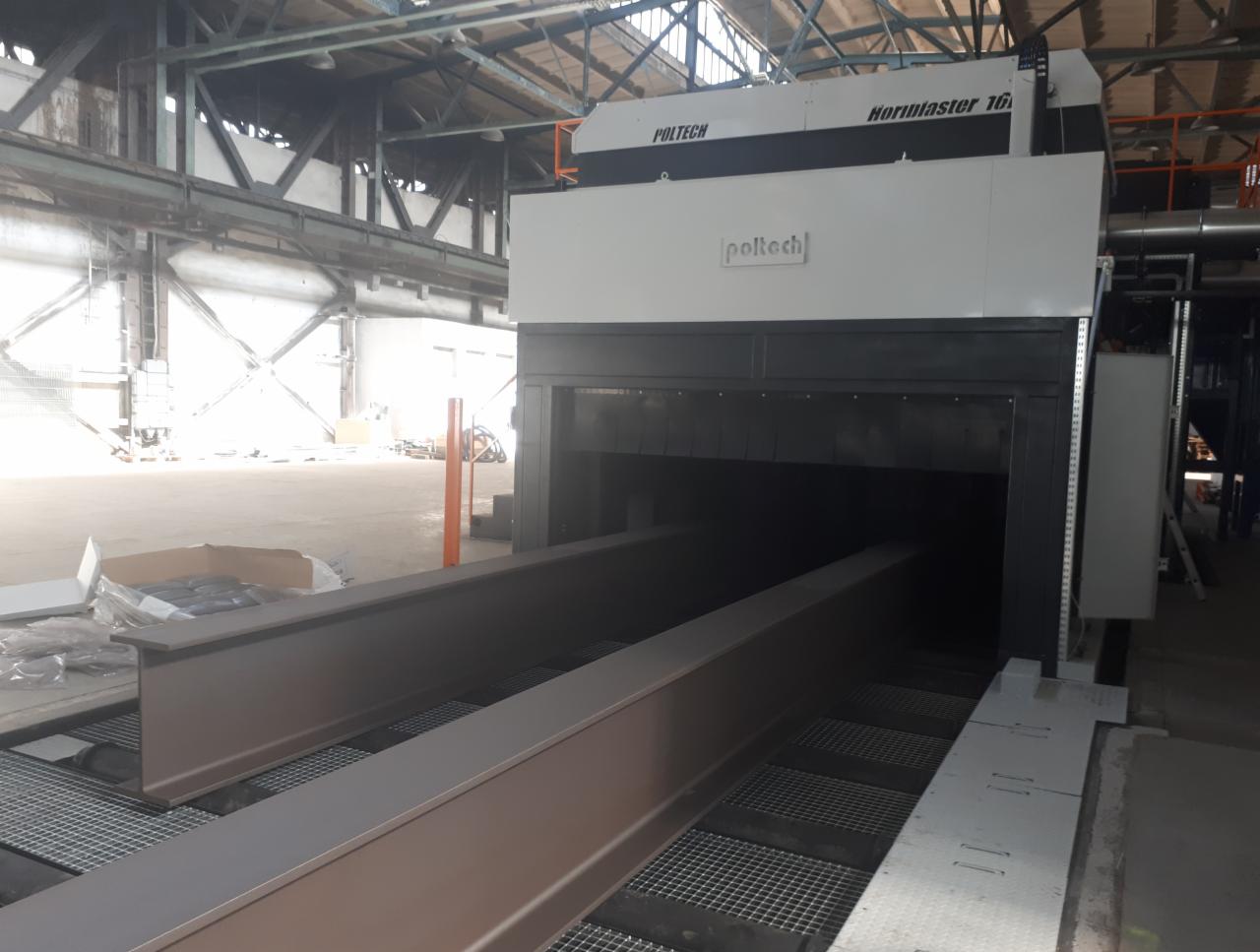 Długie, stalowe profile wchodzące do przemysłowej maszyny z napisem Poltech Hormaster 16, prawdopodobnie w trakcie procesu obróbki powierzchniowej w hali przemysłowej.