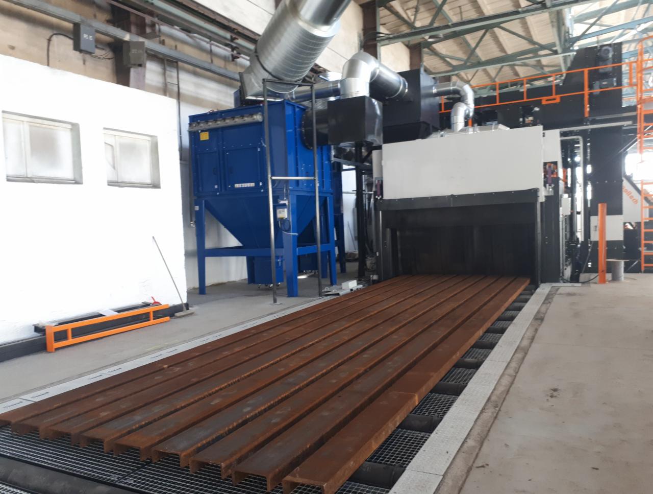 Rdzewiejące stalowe profile ułożone na podajniku przed komorą do obróbki strumieniowo-ściernej w hali przemysłowej.