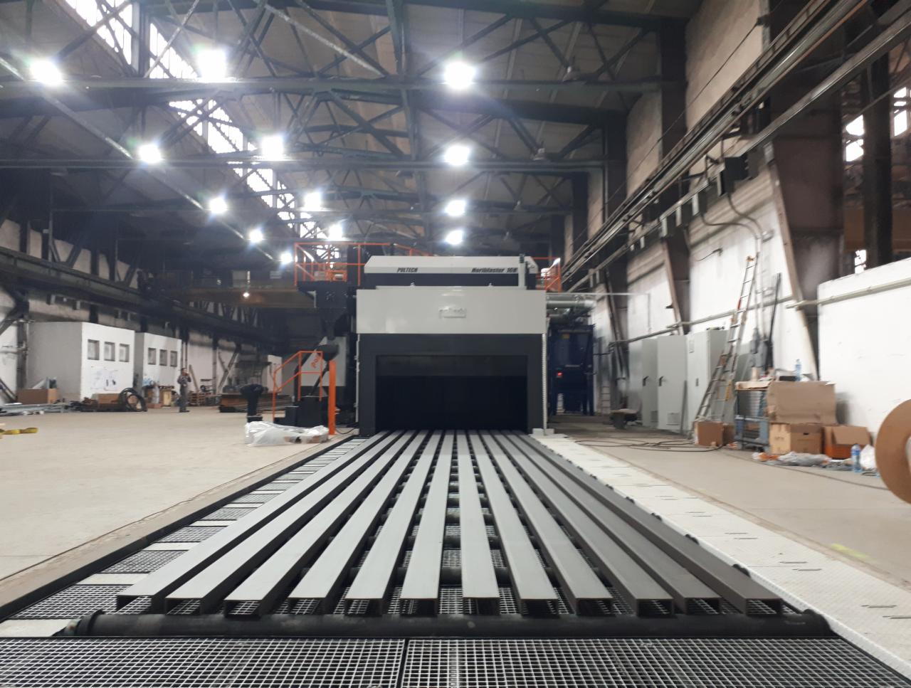 Wnętrze hali produkcyjnej z linią do obróbki metalu; widoczne profile stalowe na podajniku wjeżdżające do maszyny do śrutowania; w tle elementy konstrukcji hali i oświetlenie.