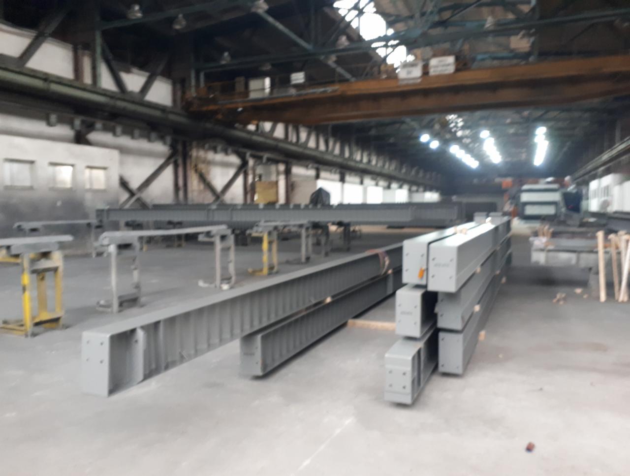 Szare, metalowe belki po obróbce, ułożone w hali produkcyjnej z suwnicą pod sufitem. Widoczne stoły warsztatowe i fragmenty innych konstrukcji.