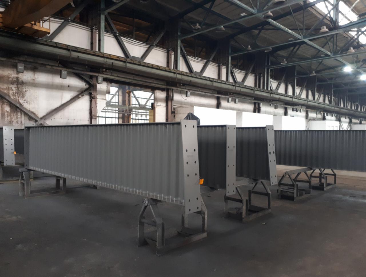 Długie, stalowe belki o przekroju skrzynkowym, świeżo pokryte szarą farbą, ustawione na metalowych kozłach w hali przemysłowej z widoczną konstrukcją dachu.