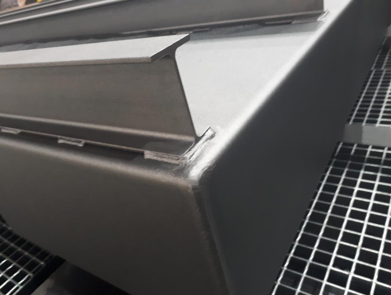 Metalowy element konstrukcyjny z widocznymi spawami i równą powierzchnią po malowaniu proszkowym, umieszczony na metalowej kratownicy.