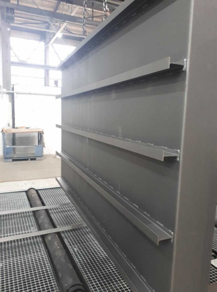 Szara, metalowa konstrukcja z trzema poziomami półek w hali przemysłowej, prawdopodobnie po procesie malowania lub śrutowania.