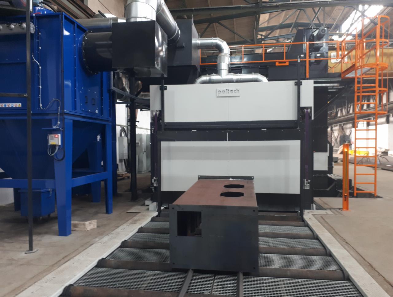 Element stalowy na taśmie transportowej przed automatyczną kabiną do śrutowania, z widocznym niebieskim separatorem pyłu i pomarańczową drabinką serwisową, w hali przemysłowej.