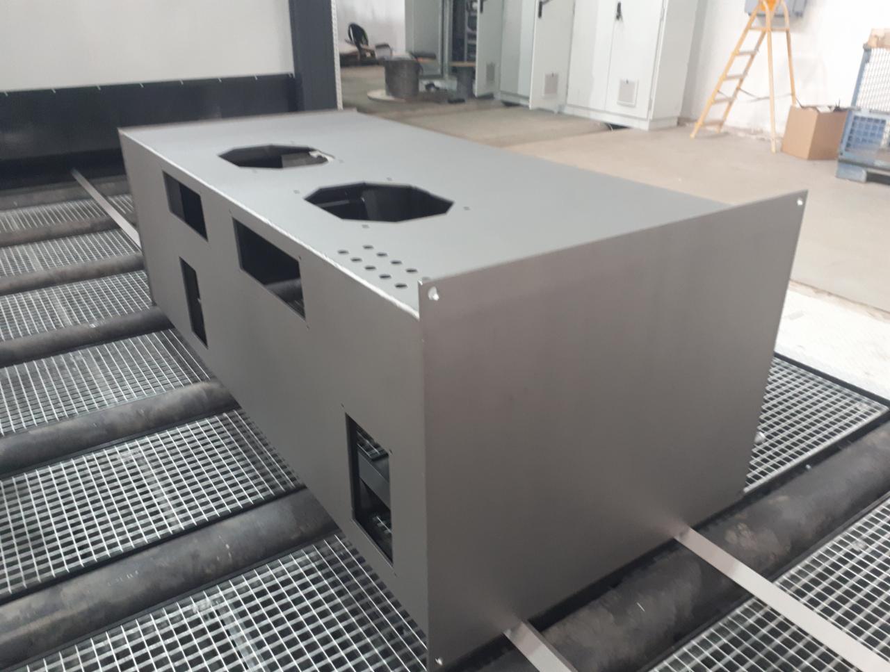 Metalowa obudowa z otworami, świeżo po malowaniu, na linii produkcyjnej z kratownicą.