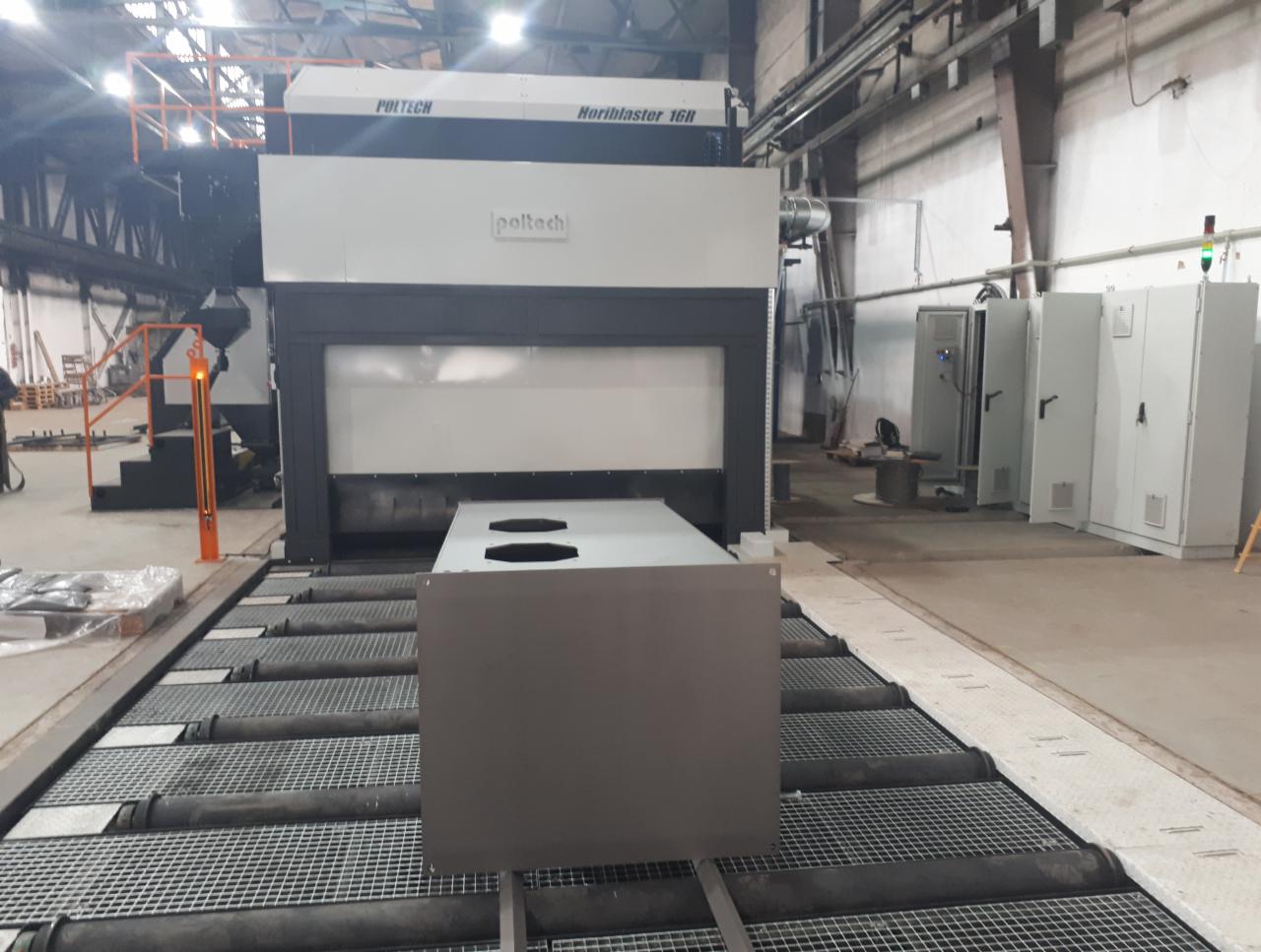 Duża maszyna do obróbki powierzchni Poltech Horiblaster 16R z elementem umieszczonym na podajniku rolkowym, widoczne szafy sterownicze w tle.