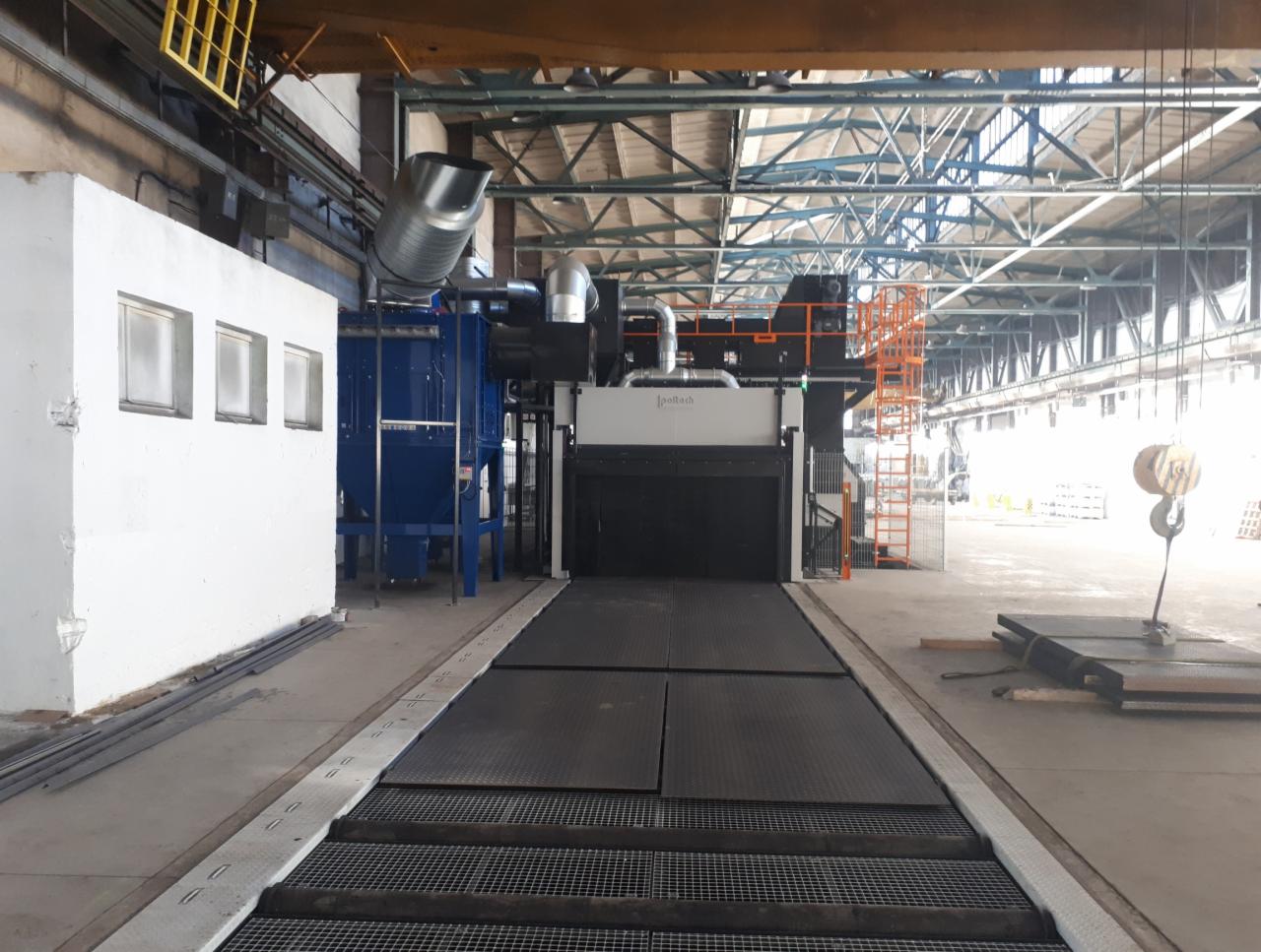 Wnętrze hali przemysłowej z dużą maszyną do obróbki powierzchni, widoczny system podajników rolkowych i instalacja odpylająca w kolorze niebieskim.