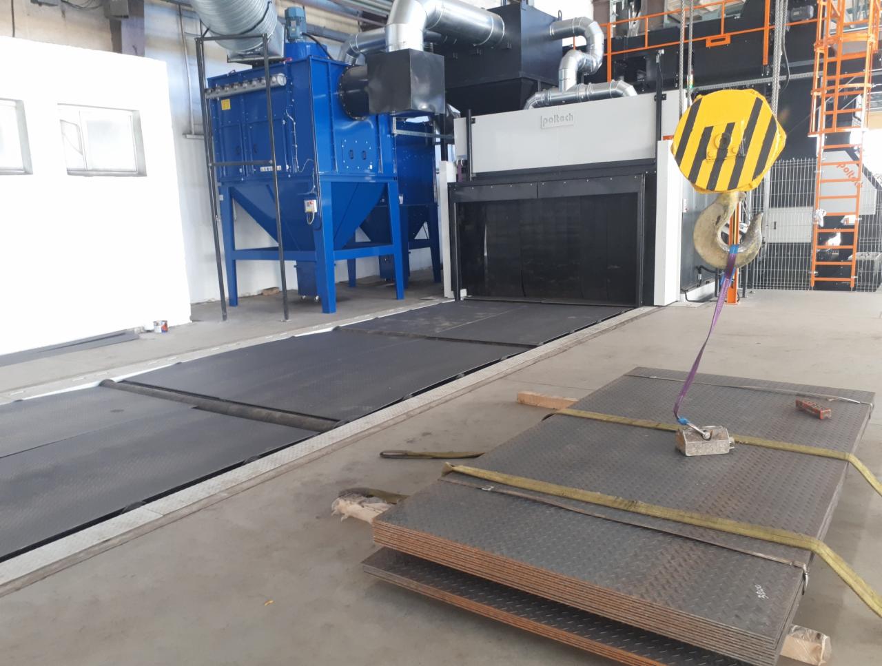 Blachy stalowe z diamentowym wzorem, podnoszone za pomocą zawiesia i magnesu, przed stanowiskiem do piaskowania w hali przemysłowej.