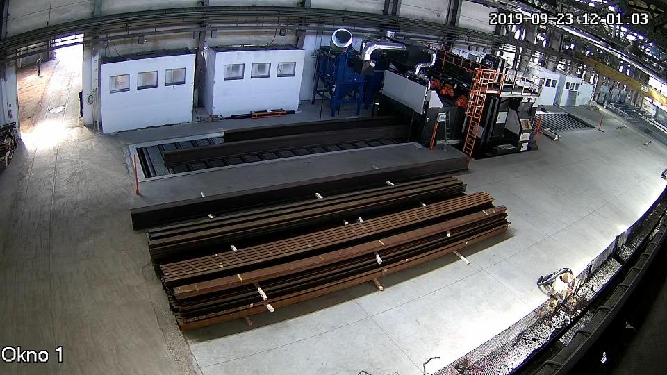 Wnętrze hali produkcyjnej z urządzeniem do obróbki strumieniowo-ściernej i stosem metalowych profili przed nim.