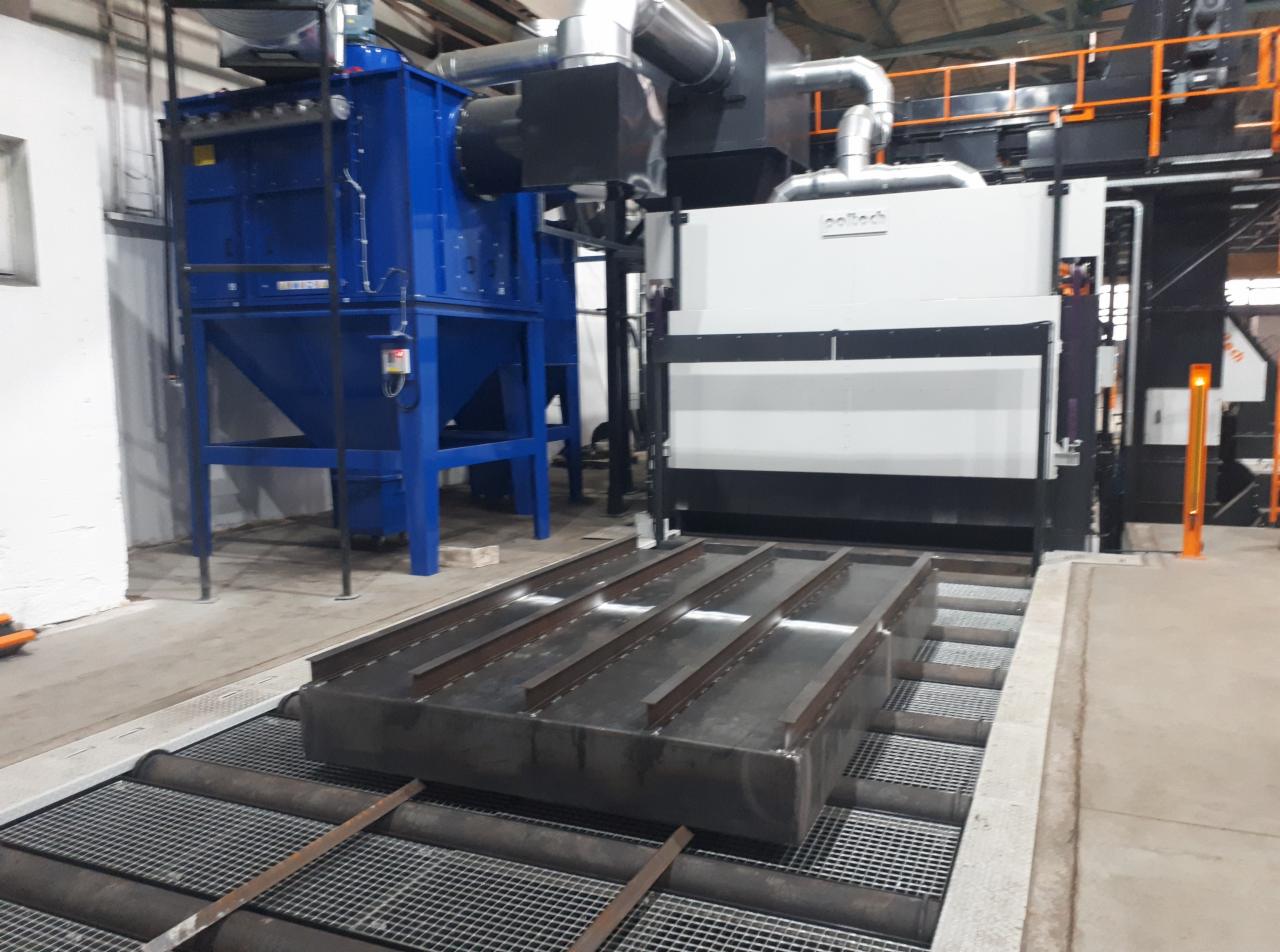 Metalowa konstrukcja na wózku transportowym przed komorą do obróbki strumieniowo-ściernej i niebieski odpylacz przemysłowy w hali.