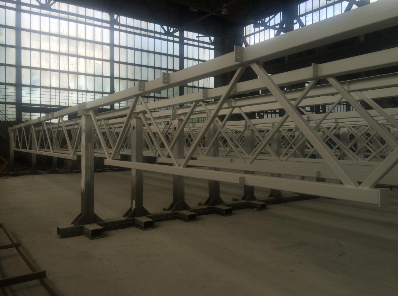 Długa, stalowa konstrukcja kratowa pomalowana proszkowo na biało, ustawiona na podporach w hali przemysłowej z dużymi oknami w tle.