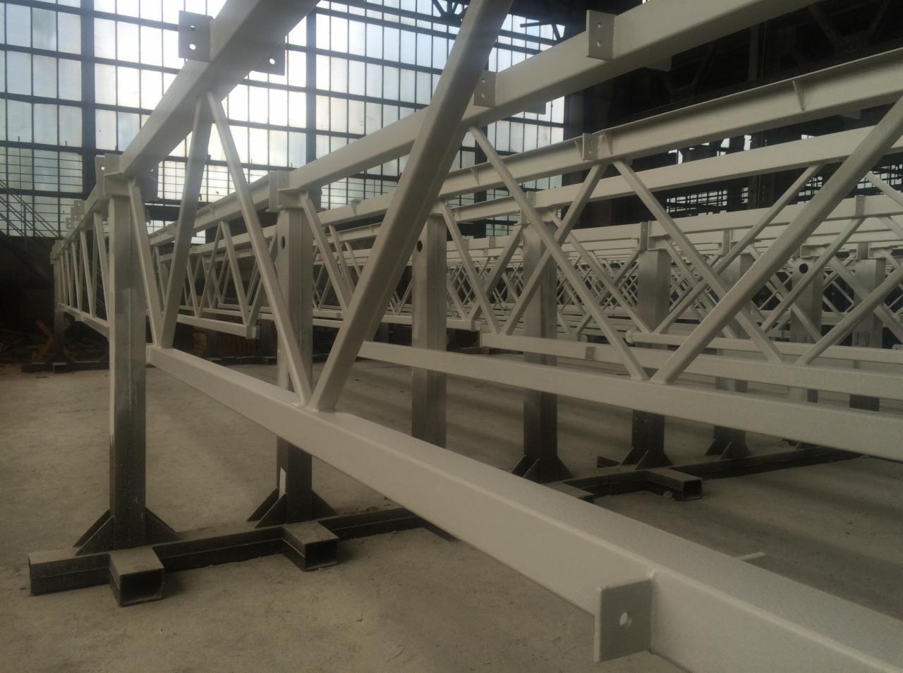 Szereg pomalowanych na biało stalowych konstrukcji kratownicowych na podporach w hali produkcyjnej, widok z niskiej perspektywy.