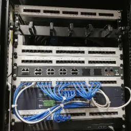 Rzędowy widok szafy rack z panelami krosowniczymi, przełącznikami sieciowymi i okablowaniem CAT6, z podświetlonymi diodami LED na urządzeniach Novus i Proxima IP PBX Server, uporządkowane...