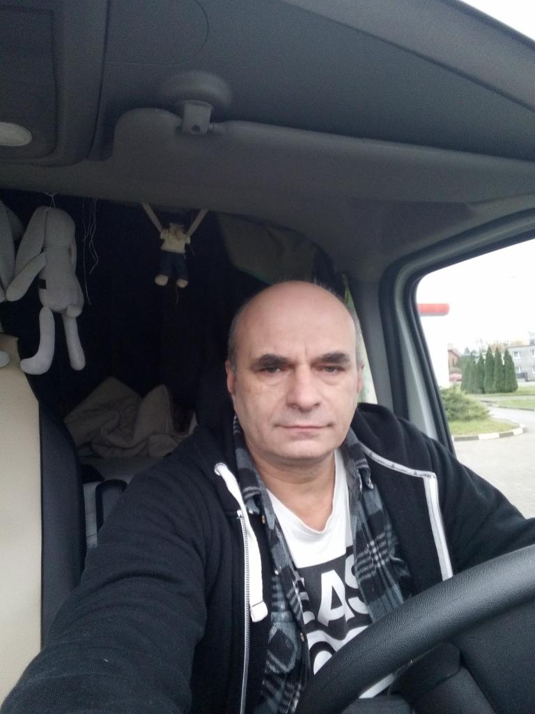 Kierowca w kabinie busa, widok z perspektywy pasażera. Zabawki wiszące nad głową, kierownica w dłoniach, budynek w tle za szybą.