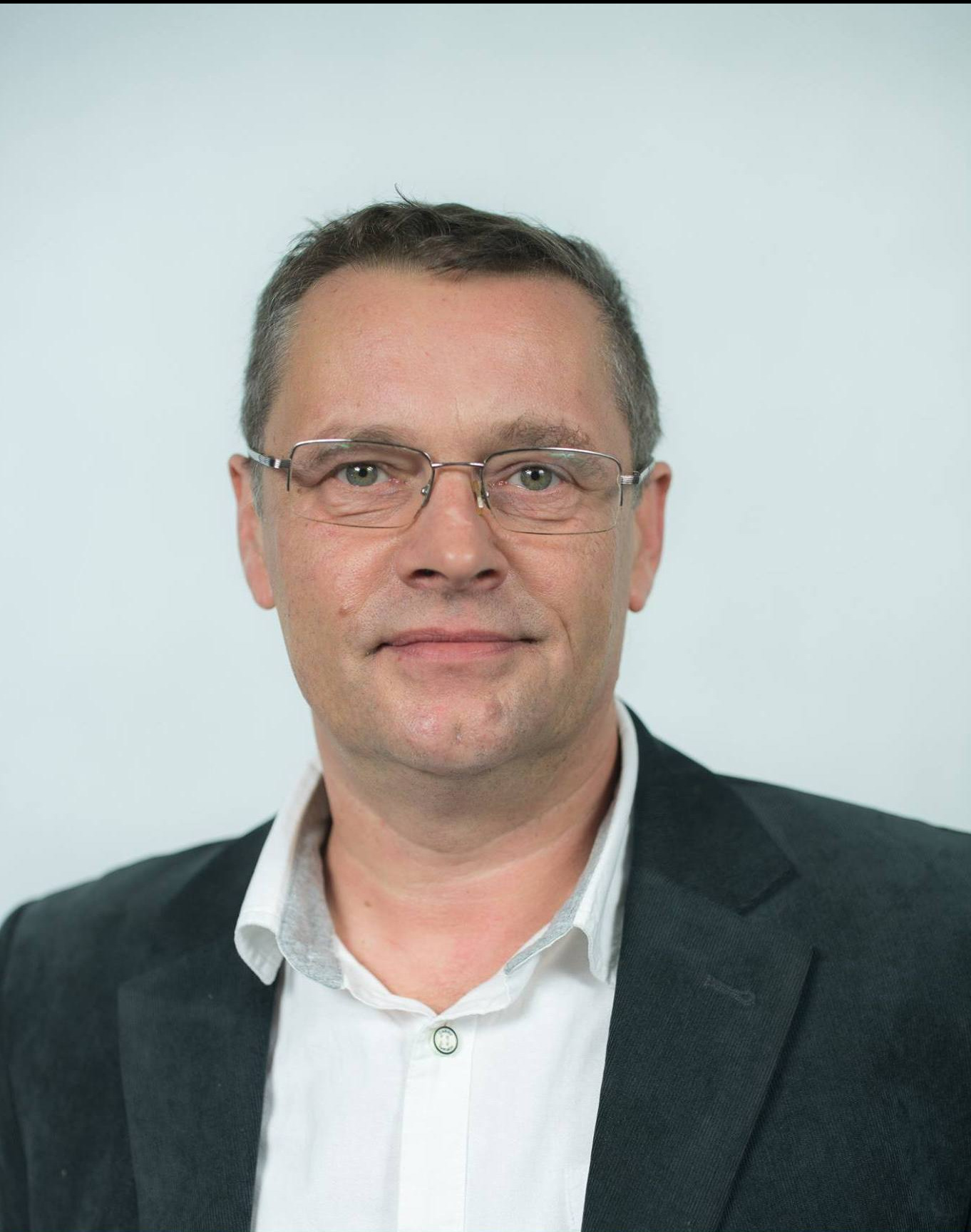 Portret mężczyzny w okularach i marynarce na jasnym tle, z delikatnym uśmiechem.