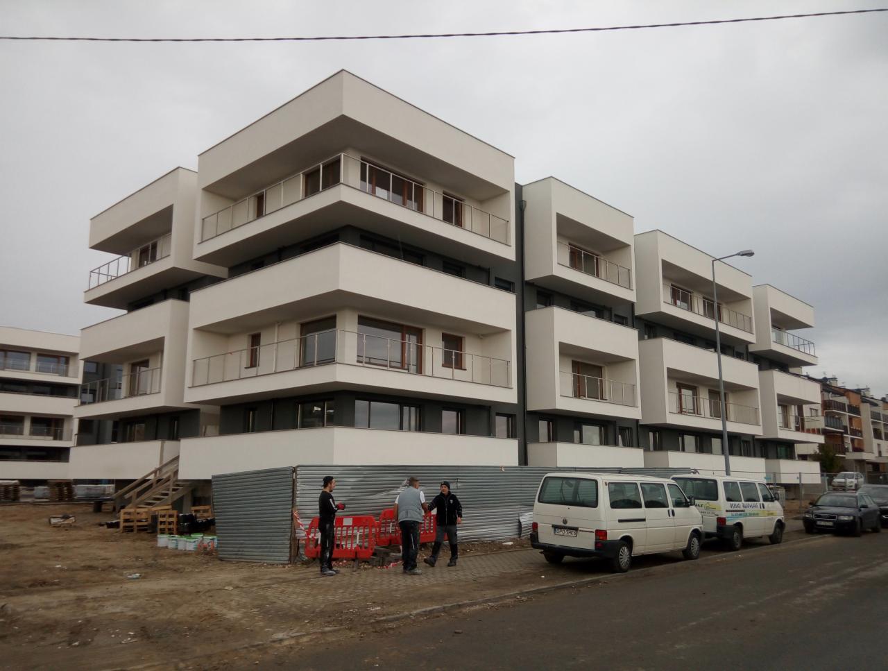 Nowoczesny budynek wielorodzinny z białymi balkonami w trakcie prac wykończeniowych, widoczne zaparkowane busy i grupa pracowników na pierwszym planie.