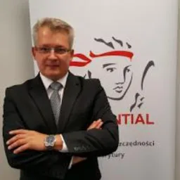 Mężczyzna w garniturze z założonymi rękami, na tle roll-upu z logo firmy ubezpieczeniowej Prudential. Mężczyzna ma okulary i zegarek na ręce.