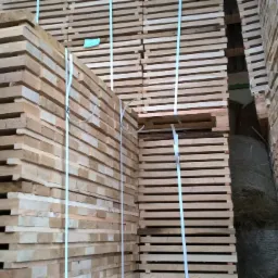 Ułożone w stosy paczki jasnej tarcicy budowlanej, zabezpieczone białymi taśmami, z widocznym fragmentem szarego materiału w tle.