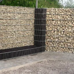 Gabionowe ogrodzenie narożne z jasnych kamieni i czarnego wypełnienia, z metalową siatką, na tle kostki brukowej i żwiru.