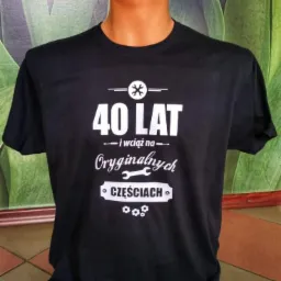 Czarna koszulka z białym nadrukiem: '40 LAT i wciąż na Oryginalnych CZĘŚCIACH', prezentowana na manekinie z zielonym, rozmytym tłem.