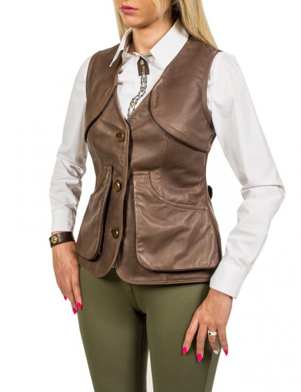 Kobieta prezentuje brązową skórzaną kamizelkę z kieszeniami, noszoną na białej koszuli z ozdobnym łańcuszkiem, w połączeniu z oliwkowymi spodniami. Widoczny skórzany zegarek na nadgarstku.