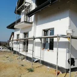 Dom jednorodzinny w trakcie prac elewacyjnych, widoczne rusztowania, ocieplenie styropianem oraz fragment dachu z czarnej dachówki. Na rusztowaniu stoi wiadro z farbą.