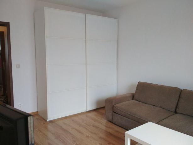 Biała szafa przesuwna z sześcioma panelami w jasnym pokoju dziennym, widoczny fragment brązowej sofy i białego stolika kawowego, podłoga drewniana.