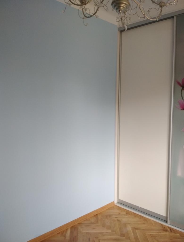 Świeżo pomalowana na jasny błękit ściana w rogu pokoju, z widoczną częścią sufitu z żyrandolem i przesuwną szafą z białą płytą oraz parkietem ułożonym w jodełkę.