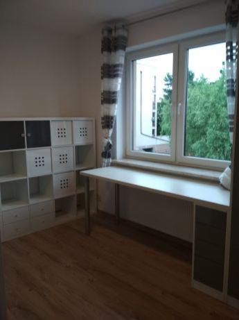 Jasne wnętrze pokoju po odświeżeniu ścian, z drewnianą podłogą, białym regałem z czarnymi akcentami i biurkiem pod oknem z widokiem na zieleń.