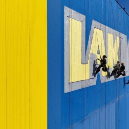 TROLL Techniki Alpinistyczne S.C. - Zespół alpinistów przemysłowych odnawia logo firmy LAMKA na elewacji budynku, widoczne liny, uprzęże i narzędzia malarskie, tło stanowi niebiesko-żółta ściana oraz oddalony krajobraz z drzewami...