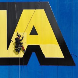 TROLL Techniki Alpinistyczne S.C. - Pracownik na linach zabezpieczających, odnawia żółty fragment logo na niebieskiej elewacji budynku przemysłowego, widoczne narzędzia i uprząż.