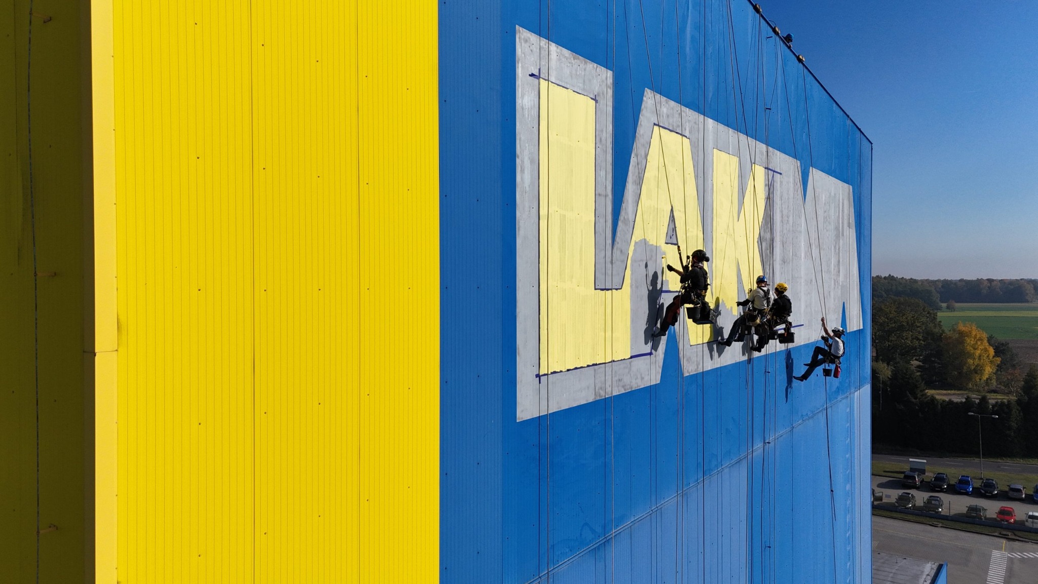 Zespół alpinistów przemysłowych odnawia logo firmy LAMKA na elewacji budynku, widoczne liny, uprzęże i narzędzia malarskie, tło stanowi niebiesko-żółta ściana oraz oddalony krajobraz z drzewami...