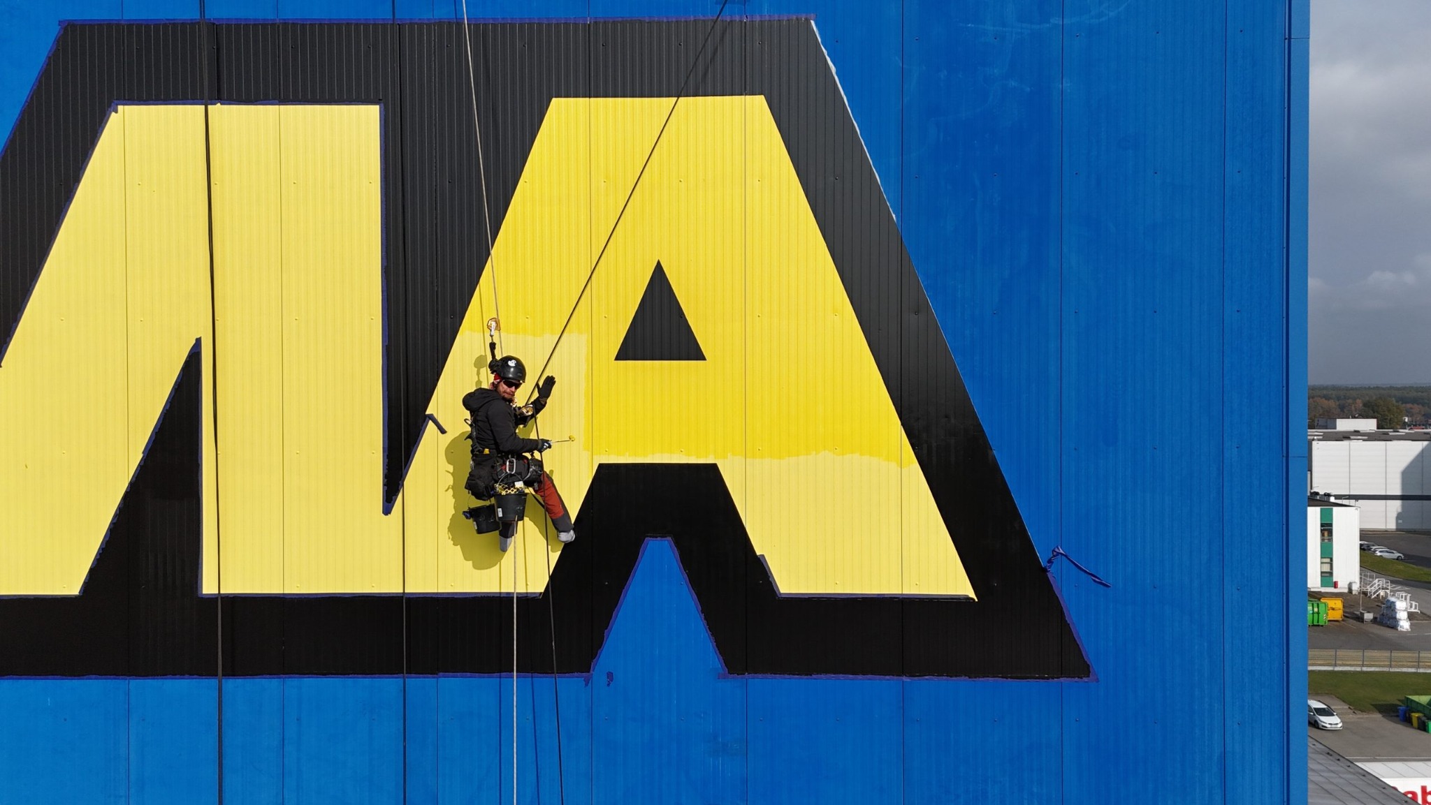 Pracownik na linach zabezpieczających, odnawia żółty fragment logo na niebieskiej elewacji budynku przemysłowego, widoczne narzędzia i uprząż.
