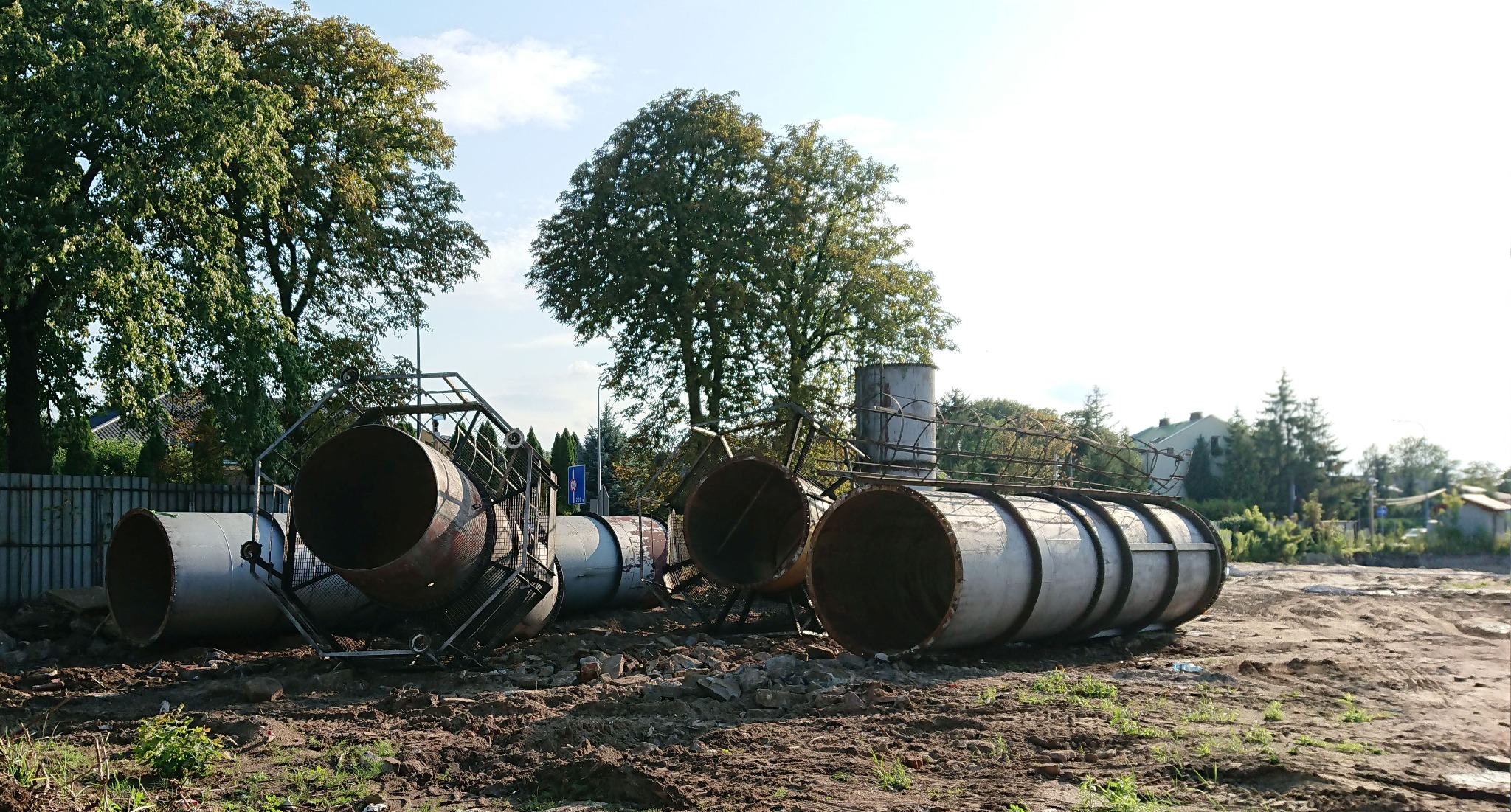 Zdemontowany, skorodowany komin przemysłowy z elementami stalowej konstrukcji leżący na ziemi, w tle drzewa i budynki.
