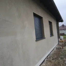 Świeżo otynkowana ściana zewnętrzna domu jednorodzinnego z zamontowanymi roletami zewnętrznymi w oknach, widok z boku w pochmurny dzień.