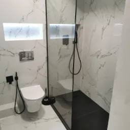 Nowoczesna łazienka z białymi marmurkowymi płytkami, czarną kabiną prysznicową typu walk-in z deszczownicą, podwieszaną toaletą i czarnymi akcentami armatury, oświetlona wnęka w ścianie.