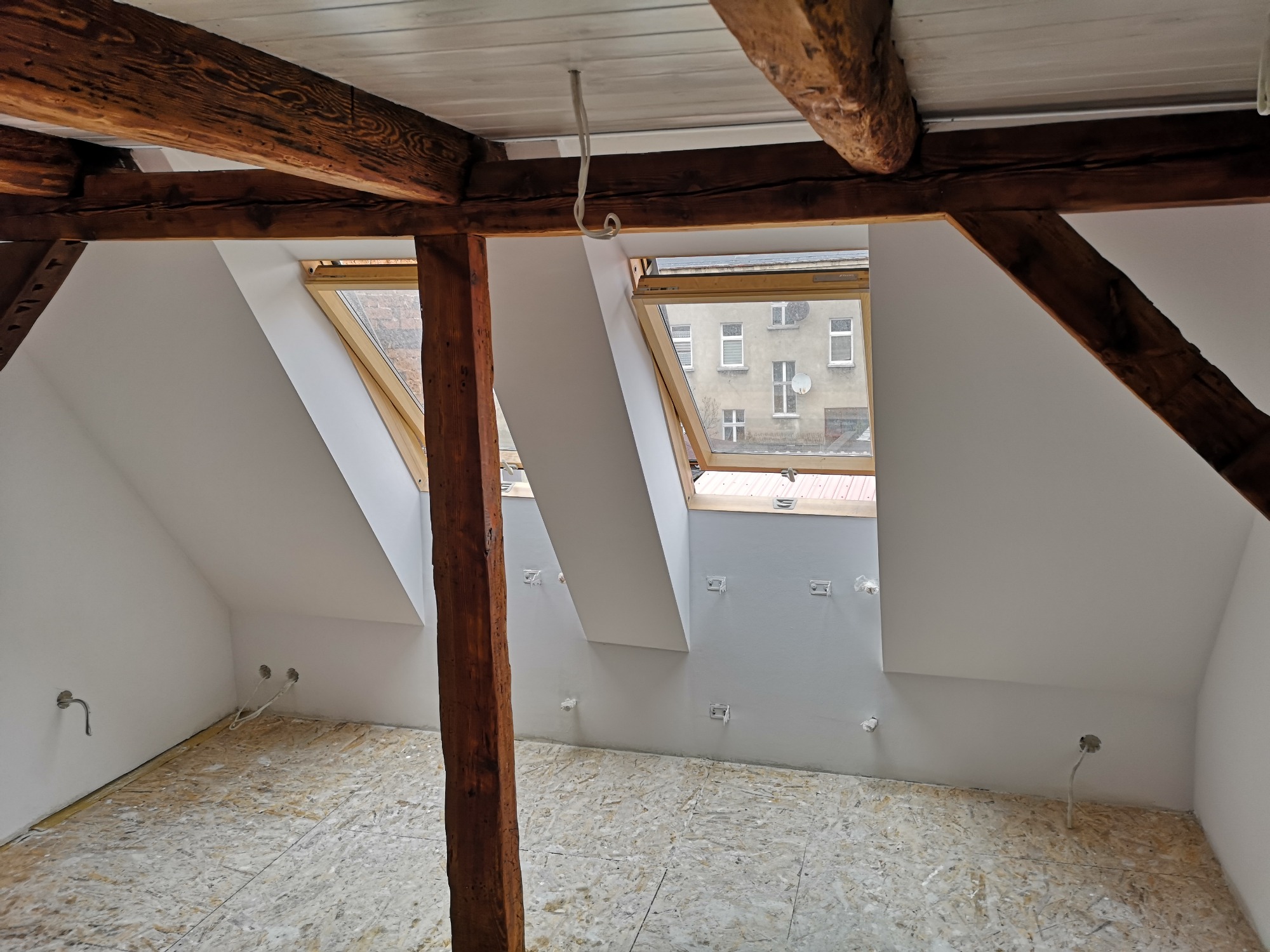 Poddasze po remoncie z drewnianymi belkami, oknami dachowymi i przygotowaną podłogą z płyt OSB oraz wyprowadzonymi instalacjami elektrycznymi.