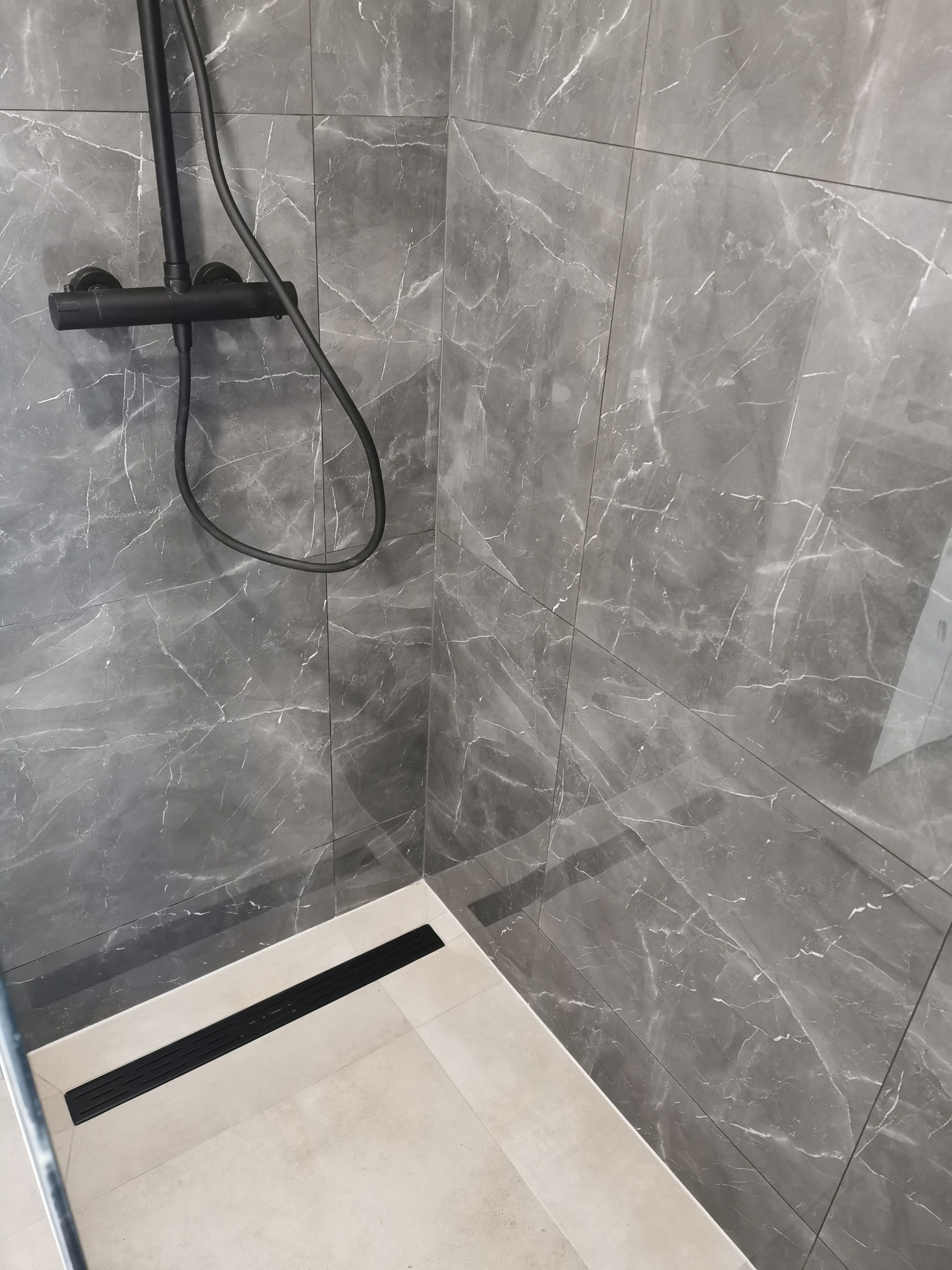 Nowoczesna kabina prysznicowa z szarymi płytkami imitującymi marmur i czarną armaturą oraz odpływem liniowym.