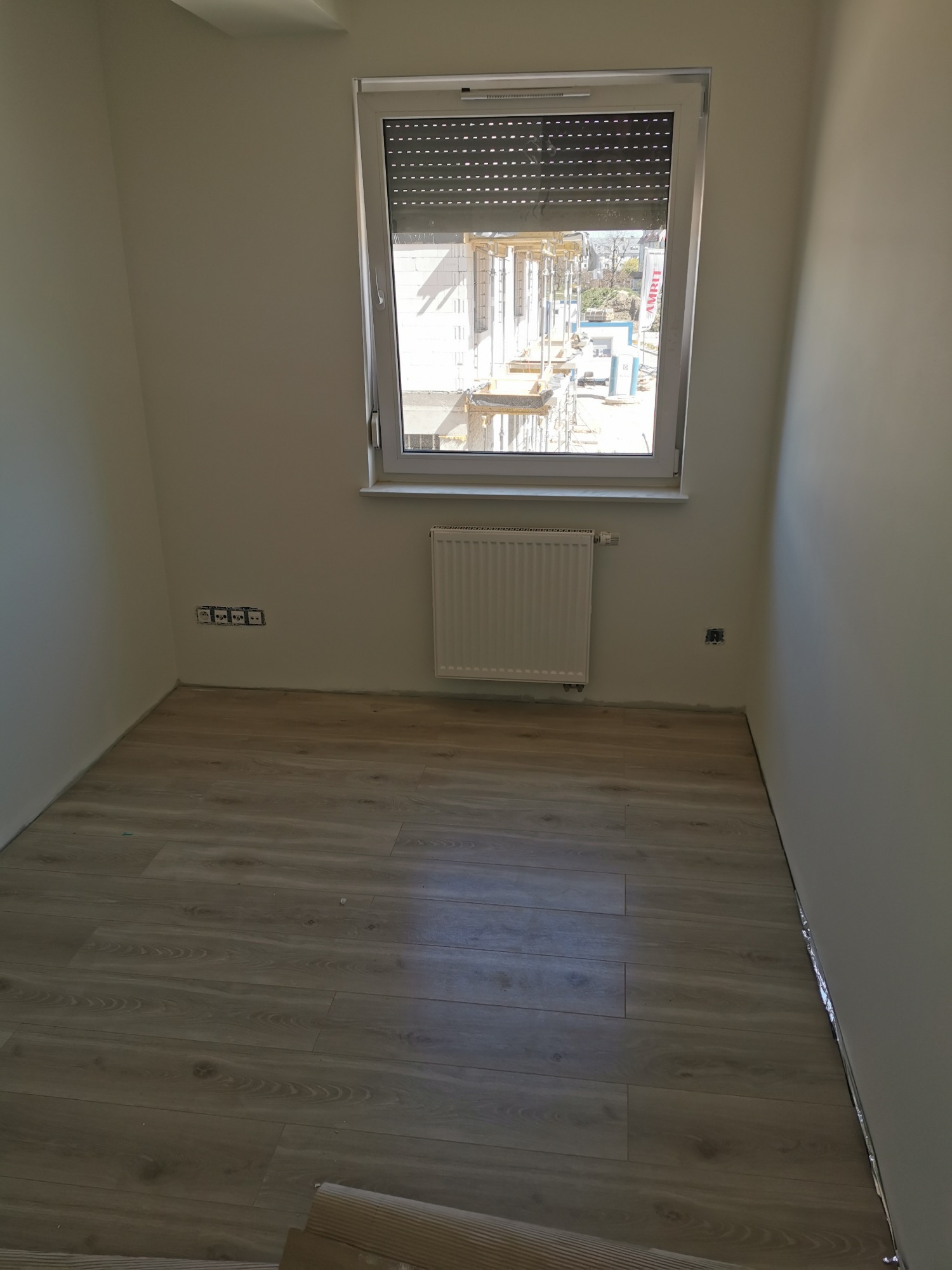 Pokój po remoncie: położona podłoga z paneli imitujących drewno, świeżo pomalowane ściany na jasny kolor, zamontowany grzejnik pod oknem z częściowo zasłoniętą roletą, widoczne gniazdka elektryczne...