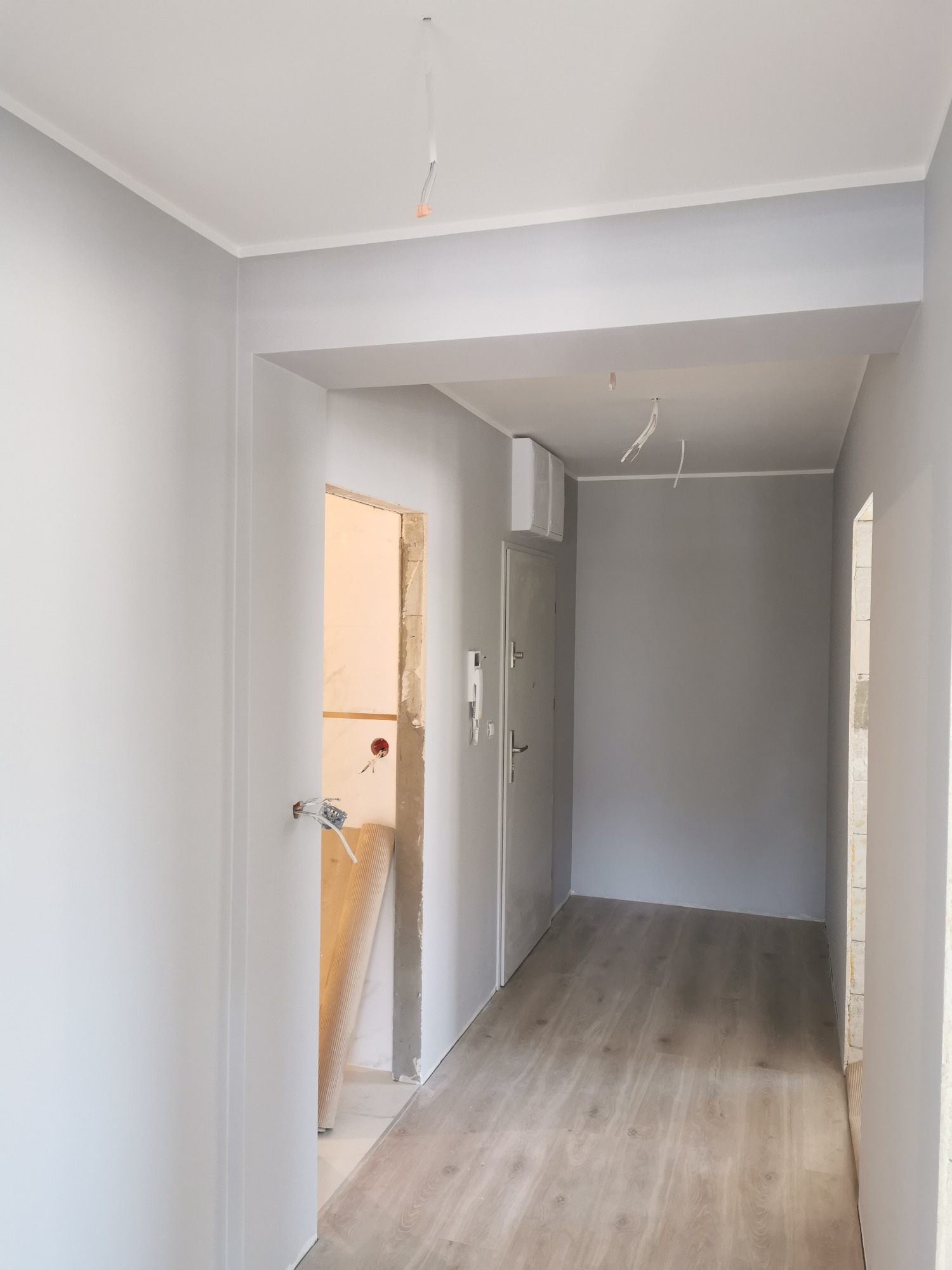 Pomieszczenie w trakcie malowania na szaro, z fragmentami ścian w stanie surowym, widoczne instalacje elektryczne i podłoga imitująca drewno.