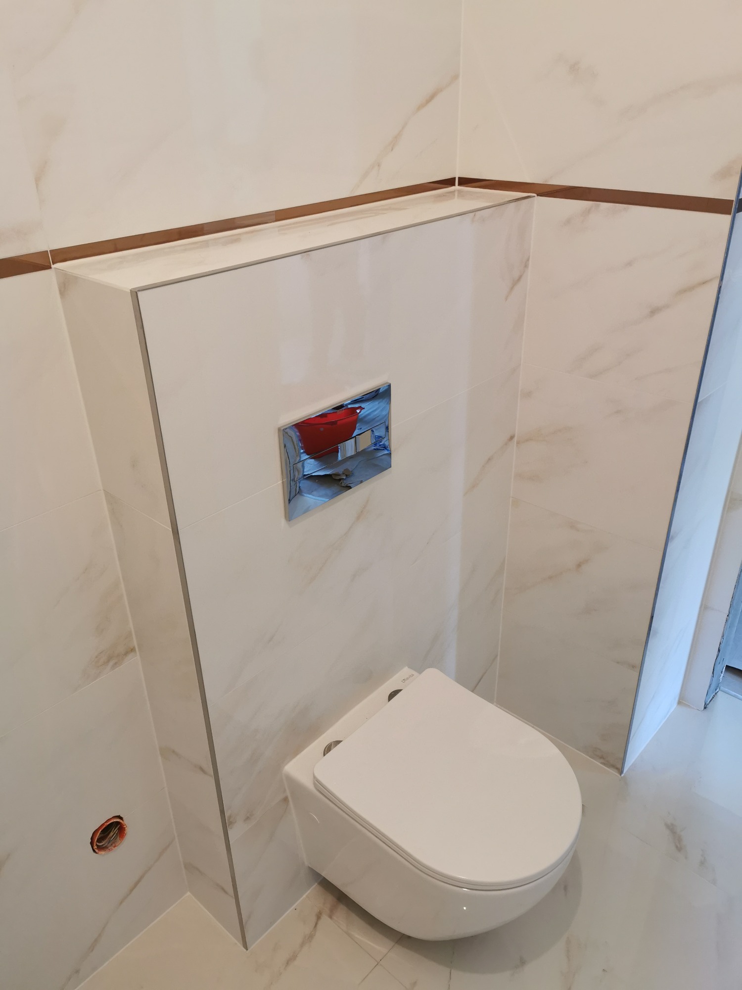 Zabudowa stelaża WC z płyt gresowych imitujących marmur, z poziomym paskiem dekoracyjnym w odcieniu brązu i chromowanym przyciskiem spłukującym.